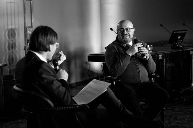 Tony D'Ambrosio seduto, microfono in mano, durante un'intervista pubblica. Fotografia in bianco e nero.