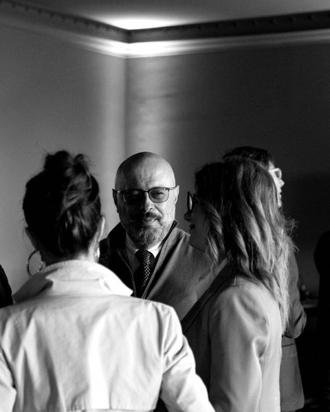 Tony D'Ambrosio ascolta, in piedi, in mezzo a tre persone di spalle durante un incontro pubblico. Fotografia in bianco e nero.